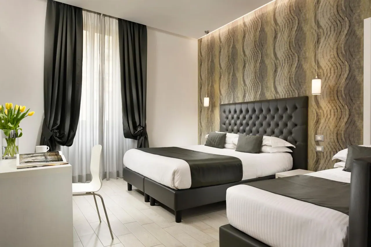 A double bed with a tufted dark headboard and a single bed share a bedroom with a white desk, a white chair and a vase of yellow tulips beside the window.