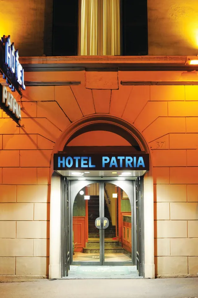 Night view of the arched entrance of Hotel Patria, with illuminated blue signs, glass doors and an orange stone facade.