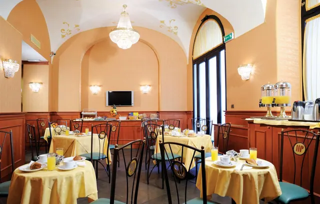 A breakfast room with yellow-clothed tables, dark metal chairs, a crystal chandelier under a vaulted ceiling, and a sideboard holding juice dispensers and coffee pots.