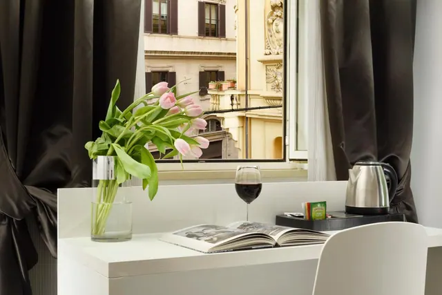 A white desk by a window holds a vase of pink tulips, a glass of red wine, an open book, a kettle and a tea tray, with a cream building facade visible outside.