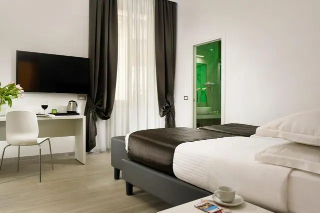 A hotel bedroom with two beds, a wall-mounted television, a desk with a chair and a kettle, and a green-lit bathroom visible through an open door.