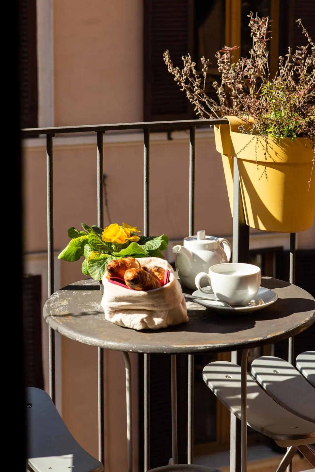 A round metal table on a small balcony holds a teapot, a cup and saucer, and a bag of croissants beside a yellow flowering plant, with a building wall behind.
