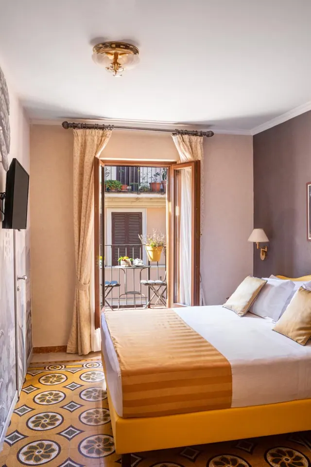 A bedroom with a double bed, a wall-mounted television, and open French doors onto a small balcony with a bistro table and chairs.