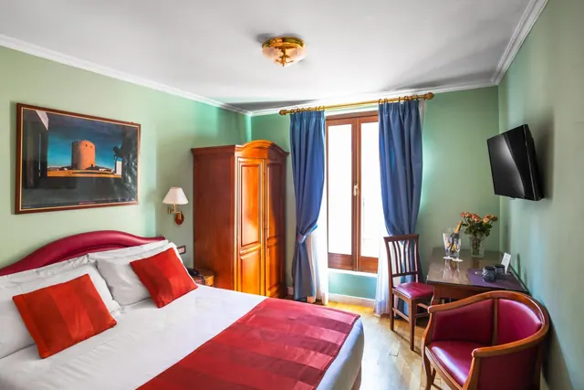A double bed with red accents sits in a green-walled bedroom beside a wooden wardrobe, a wall-mounted television and a small writing desk with chairs.