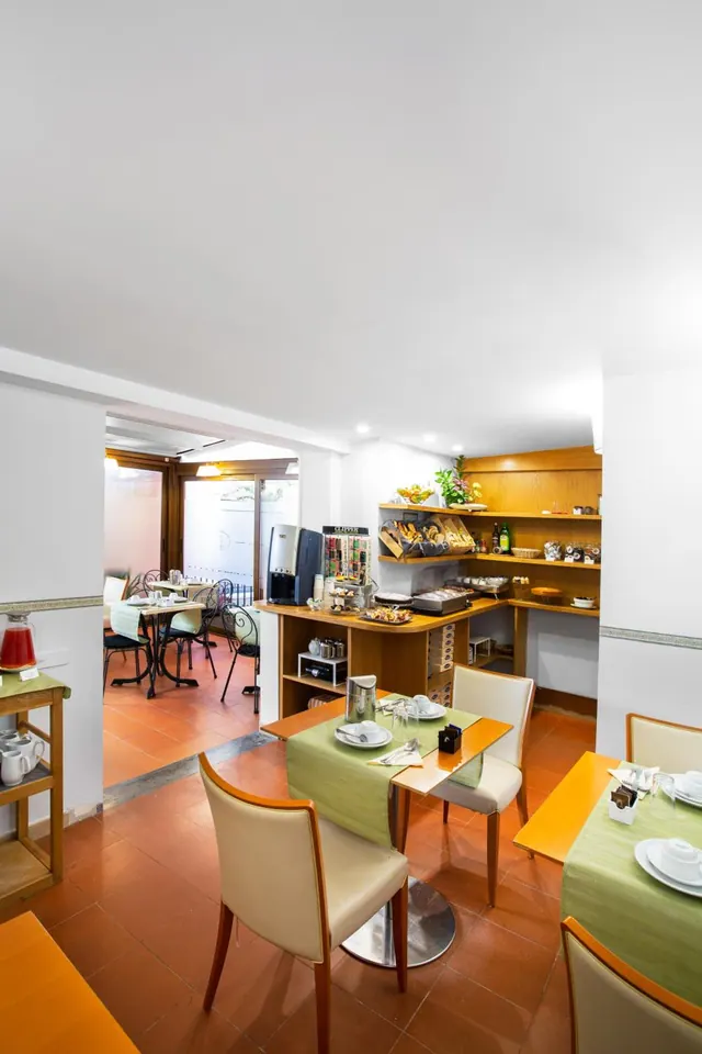 A breakfast room with set tables, green runners and cream chairs, a wooden buffet counter with pastries and a coffee machine, and a further seating area visible through the doorway.