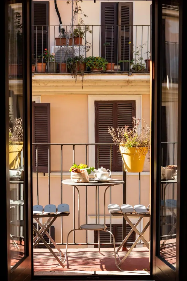 A small round table with a teapot, cup and bread on a sunlit balcony, seen through open French doors, with a yellow planter and a neighbouring building facade behind.