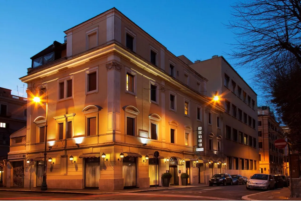Exterior of a lit multi-storey hotel building at dusk, with a vertical hotel sign, street lamps and parked cars along the road.
