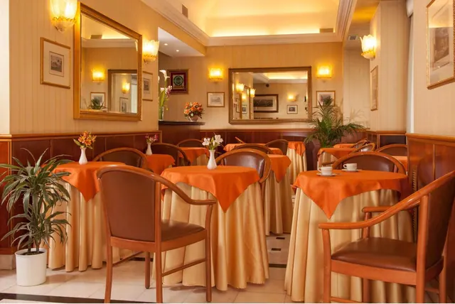 A dining room with round tables covered in orange and gold cloths, wooden chairs with brown leather seats, mirrors and wall lights on cream walls, and a potted plant at left.