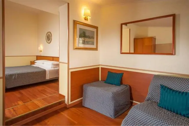 A seating area with a grey sofa and ottoman with teal cushions, a framed picture and mirror on the wall, and a bed visible through the doorway beyond.