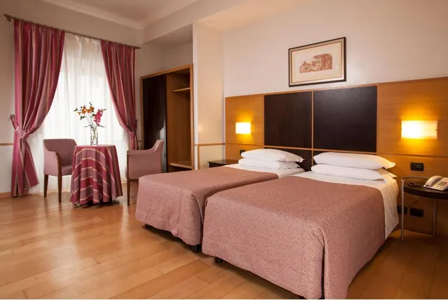 A bedroom with two twin beds, pink striped curtains, two armchairs and a small round table with flowers.