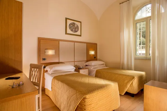 A bedroom with two single beds with gold quilts, a wooden headboard, a desk with a phone and glasses, and an arched window showing trees outside.