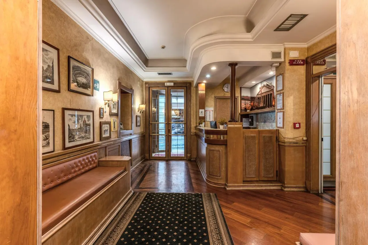 A hotel lobby with a tufted leather bench along the left wall, framed prints of Rome, a wooden reception desk and a patterned runner rug on a wooden floor.