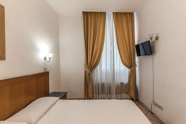 A bedroom with a double bed under a wooden headboard, a window with brown curtains and sheer net curtains, and a wall-mounted television beside wall lights.