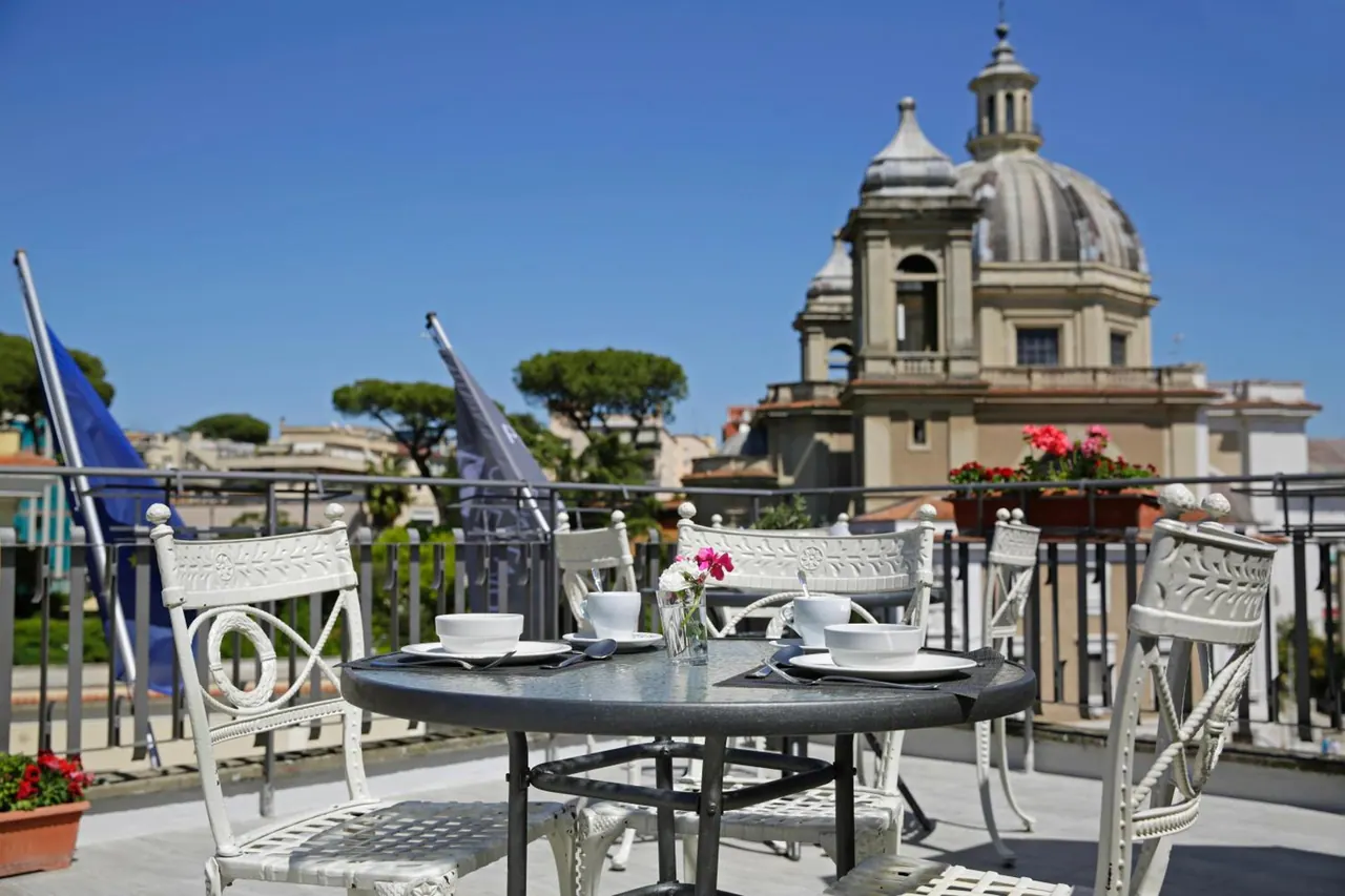 A set round table with cups, saucers and a small flower vase sits on a rooftop terrace, with a domed church and rooftops in the background.