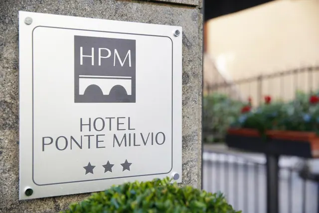 A metal plaque on a stone wall shows the HPM logo and the name Hotel Ponte Milvio, beside a railing and flower boxes.