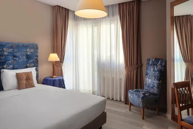 A bedroom with a made double bed, a blue upholstered armchair, a bedside table with a lamp, and sheer curtains over a tall window.