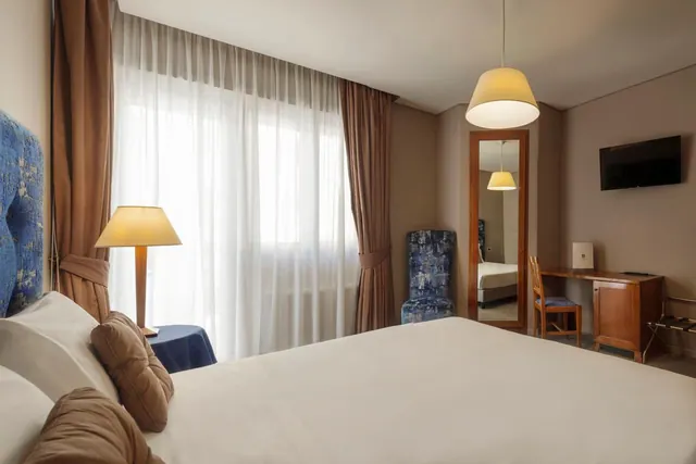 A hotel bedroom with a large bed, a table lamp beside sheer-curtained windows, a blue armchair, a full-length mirror, a wooden desk and a wall-mounted television.