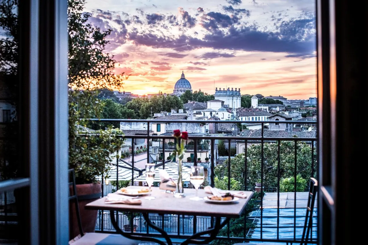 A balcony table is set with wine glasses, plates and a red rose, with a view over Rome's rooftops and a domed basilica at sunset.
