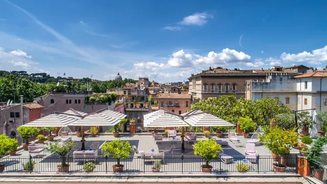 A rooftop terrace with striped umbrellas, lounge chairs and potted citrus trees overlooks Rome's rooftops and a distant dome under a blue sky.
