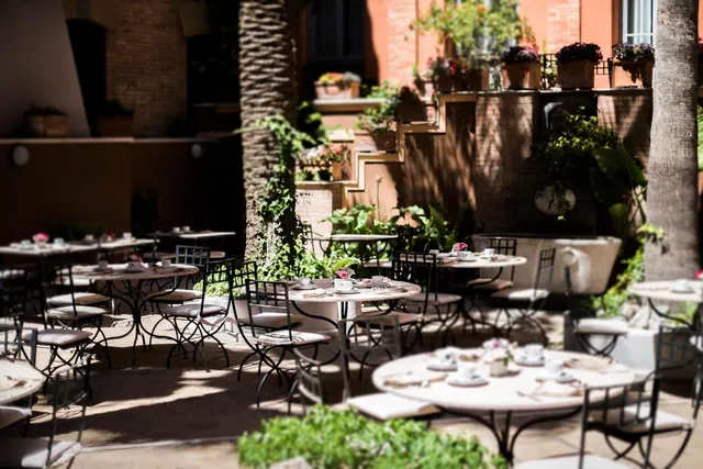 An outdoor courtyard dining area with round tables set with cups and saucers, metal chairs with cushions, palm trees and potted plants beside a terracotta building.