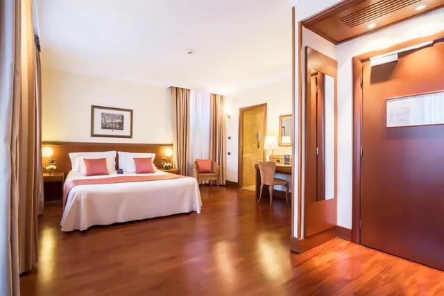 A double bed with pink cushions, an armchair and a desk with a chair stand in a hotel bedroom with a wooden floor.