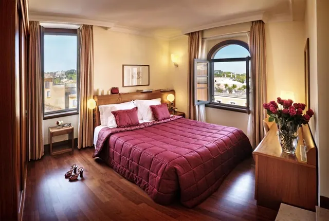 A bedroom with a double bed under a magenta quilted cover, two windows looking out over rooftops and trees, and a wooden dresser holding a vase of red roses.