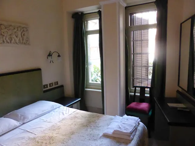 A double bed with a green headboard and folded towels on the quilt, with two tall windows and green curtains beside it.