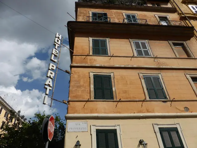 Looking up at a multi-storey ochre building with green shutters, a vertical Hotel Prati sign on its side and a stop sign at street level.