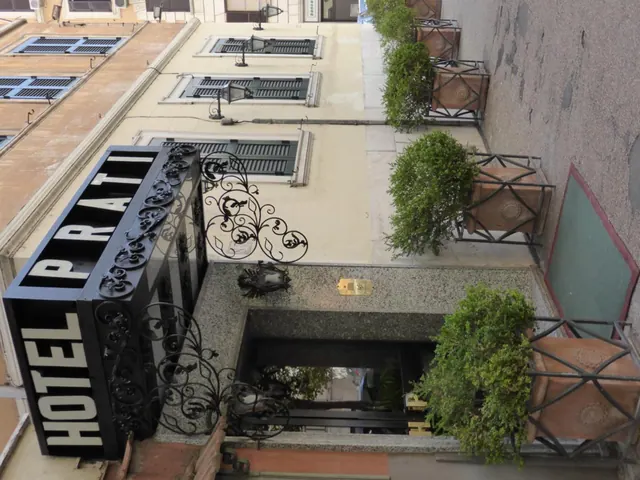 Looking down at the entrance of Hotel Prati, with a black sign, ornate wrought iron decoration, potted shrubs in terracotta planters and shuttered windows on the facade.