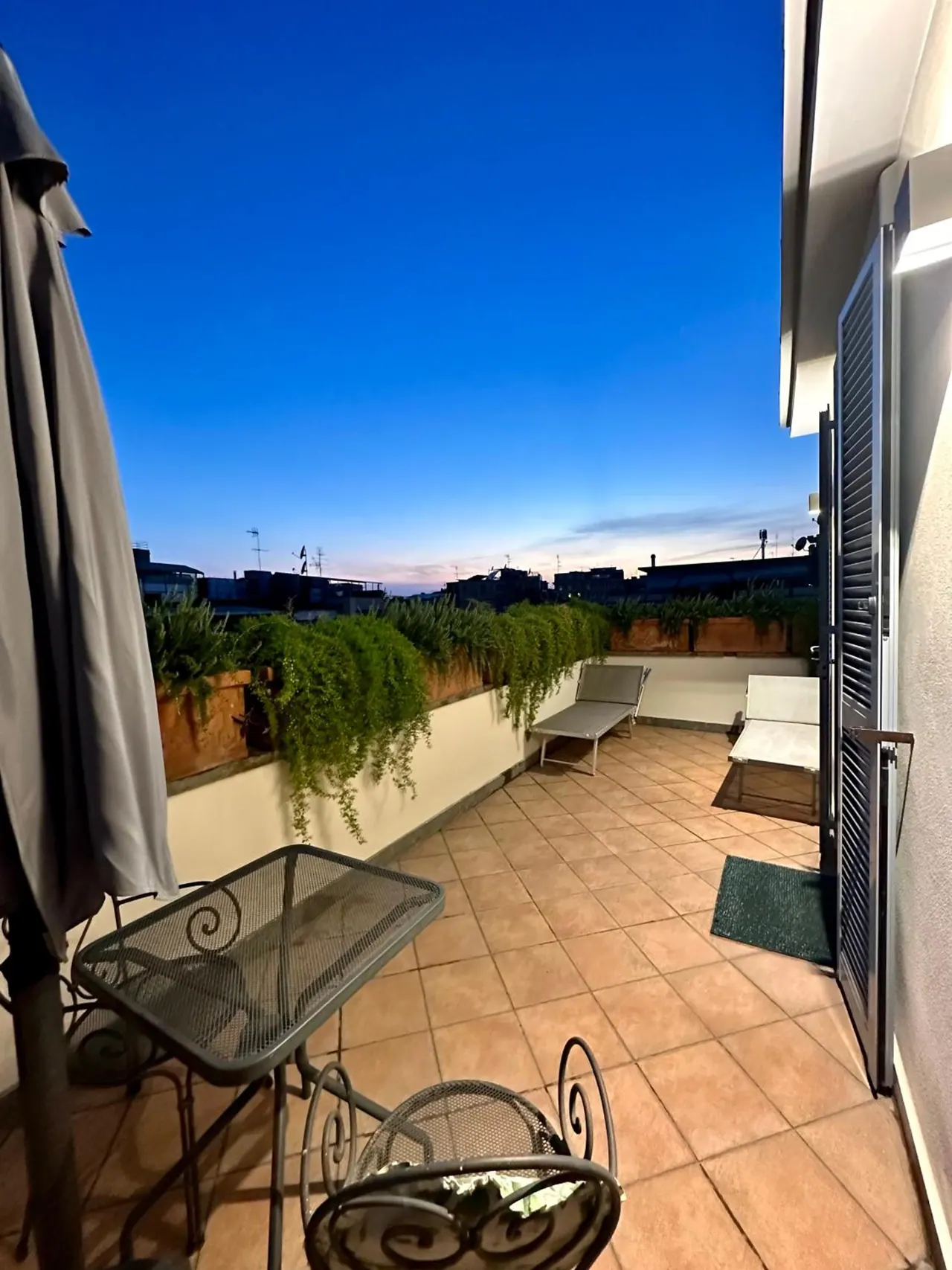 A rooftop terrace at dusk with a metal table and chairs, two sun loungers, planters with greenery, and a view over city rooftops under a blue evening sky.