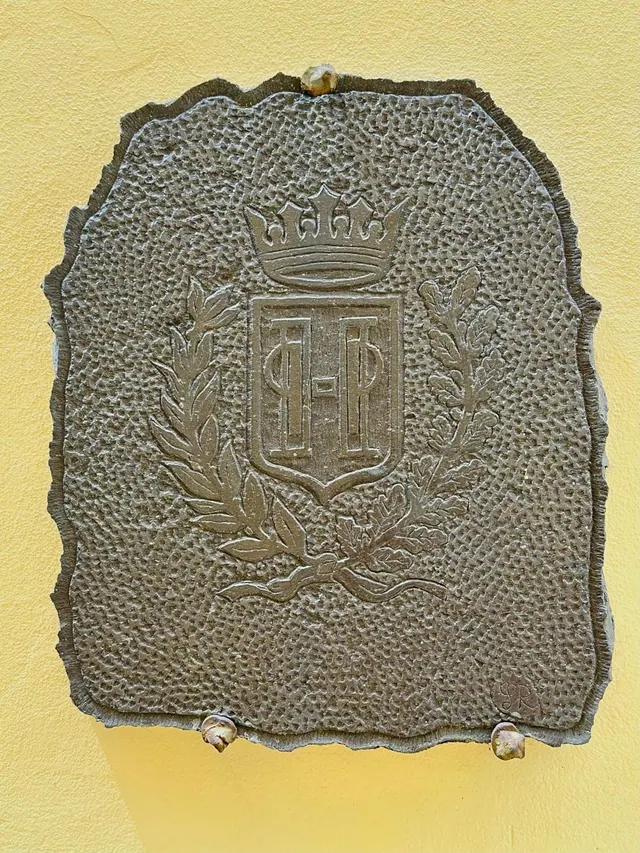 A textured metal coat of arms with a crown, laurel and oak branches is mounted on a yellow wall.
