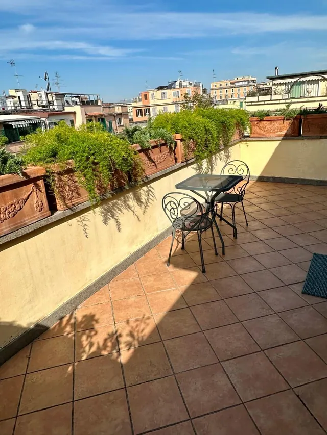 A sunny rooftop terrace with terracotta floor tiles, planters, a glass-topped table with two wrought iron chairs, and views of surrounding city rooftops.