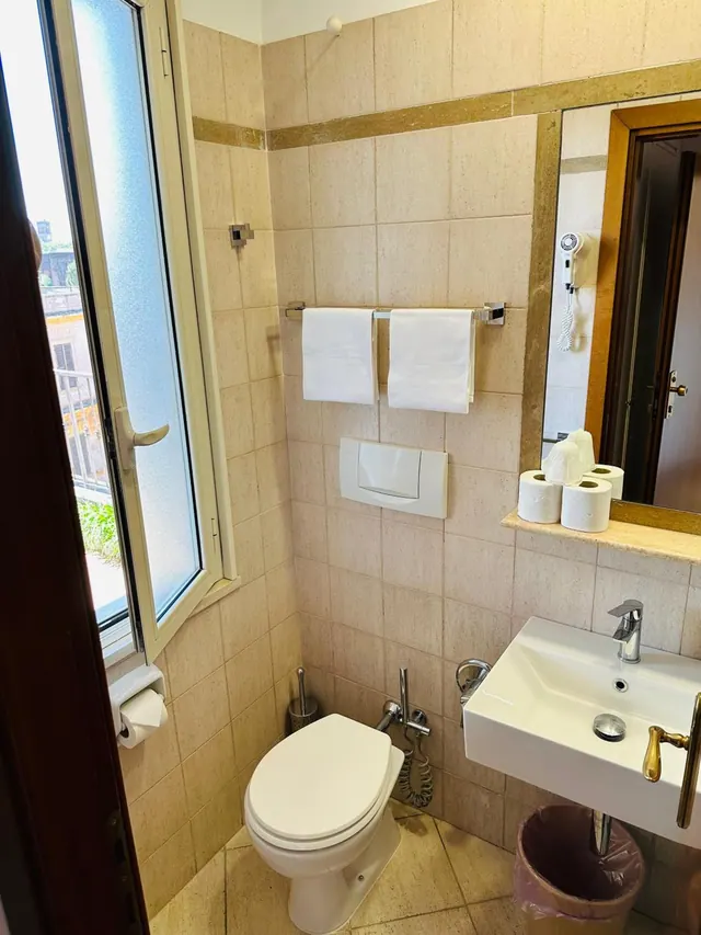 A bathroom with beige tiled walls, a toilet with a hand bidet spray, a wall-mounted sink, and a window showing neighbouring buildings.
