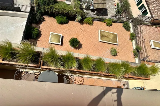 Looking down from a balcony onto a tiled courtyard with raised planters of grasses and shrubs, a sunken rectangular water feature, and a wrought iron table and chairs at the lower left.