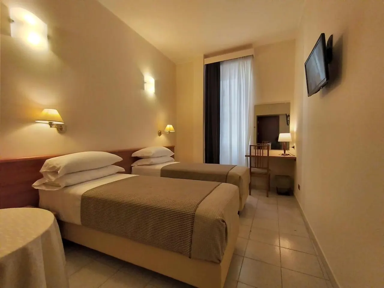 A hotel bedroom with two single beds with grey-brown throws, wall lamps, a wall-mounted television, and a small desk with a chair beside a curtained window.