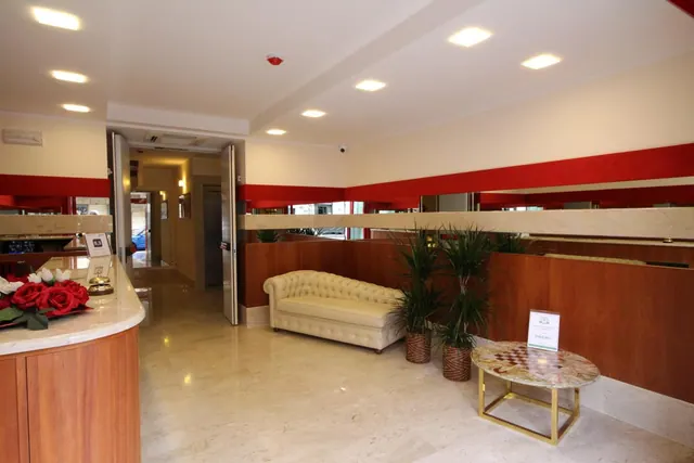 The lobby has a curved reception desk with red flowers, a cream tufted sofa, potted plants and a round marble-topped table on a pale marble floor.