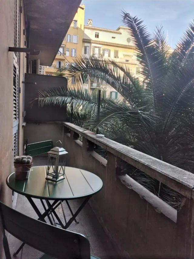 A small balcony with a round green metal table, two green chairs, a lantern and a potted cactus, with a palm tree and yellow buildings beyond the railing.