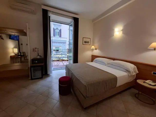 A made double bed with white pillows and a brown throw sits in a hotel room, with glass doors open onto a small balcony that has a table and chairs.