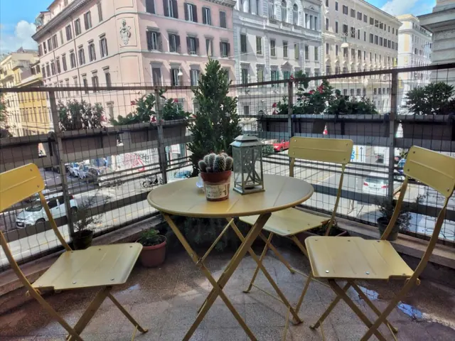 A balcony with a yellow metal bistro table, three folding chairs, a cactus pot and a lantern, overlooking a street with parked cars and pale apartment buildings.
