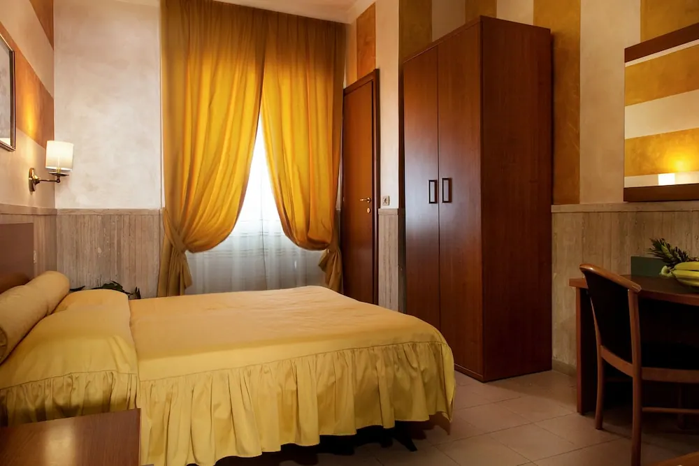 A bedroom with a yellow bedspread on a double bed, a wooden wardrobe, a desk with a bowl of bananas, and a window with yellow curtains.