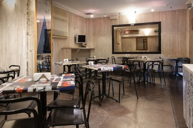 A dining room with black metal chairs and tables topped with newspaper-print covers, a wall-mounted television and a large framed mirror.