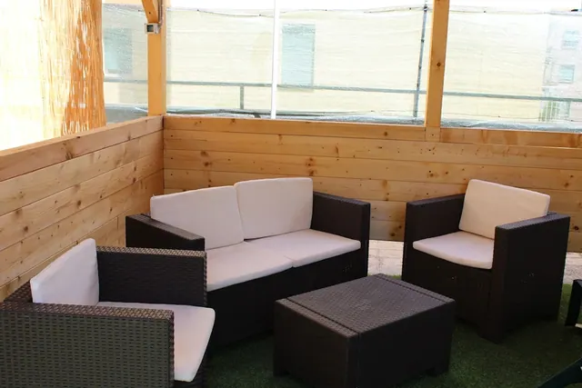 A covered wooden terrace with a dark wicker sofa and armchairs with white cushions, a low table and an artificial grass floor, with windows looking onto a building through a mesh screen.