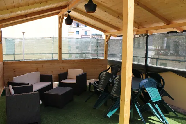 A covered wooden terrace with wicker sofas, a coffee table, and green plastic chairs around a table on artificial turf, with shade netting along the railings.