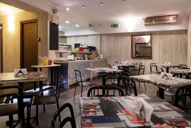 A breakfast dining room with tables covered in London-themed cloths and folded napkins, and a buffet counter at the back holding juice, fruit, plates and a coffee machine.