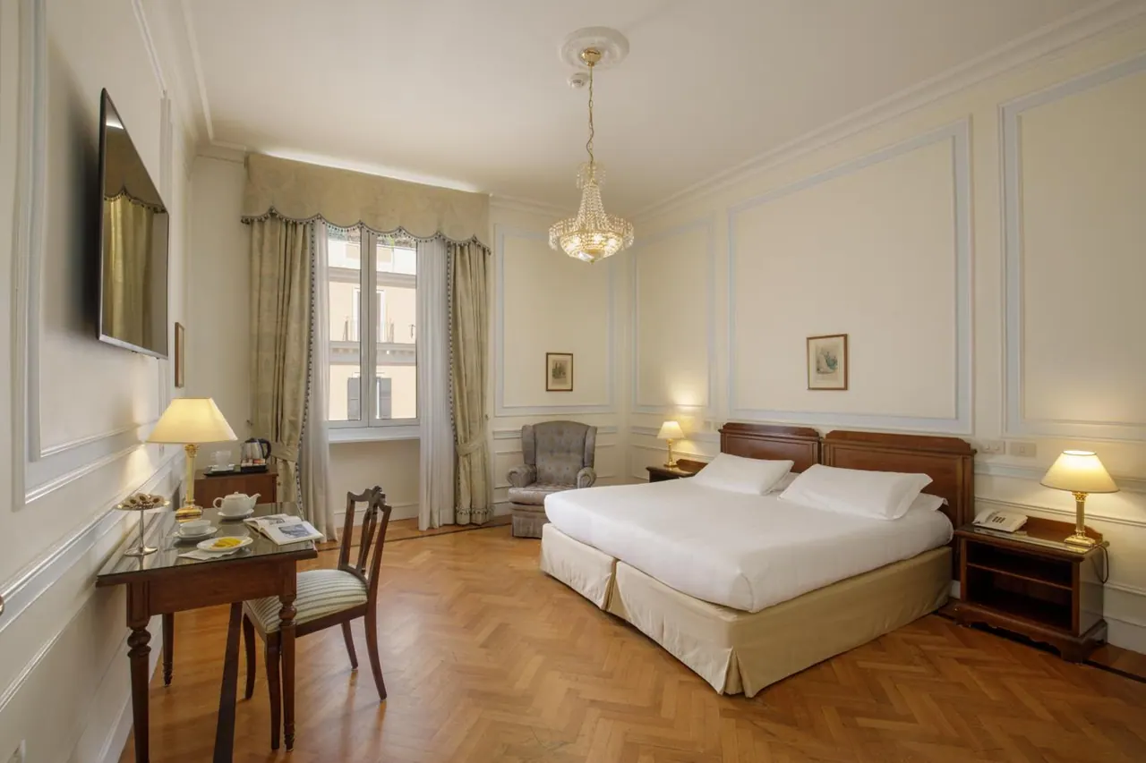 A double bed with white linens sits beside a wooden writing desk and an armchair in a room with a crystal chandelier and a tall window.