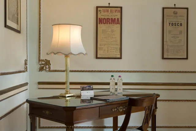 A wooden desk with a glass top holds a lit table lamp, two water bottles and a folder, with a chair beside it and framed opera posters on a cream wall with gold moulding.