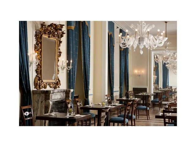 A dining room with blue curtains, a gilded mirror, a marble fireplace, crystal chandeliers and set tables with blue upholstered chairs.