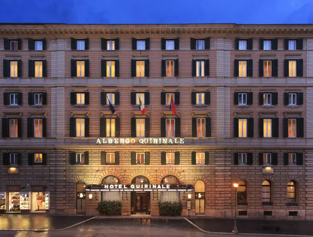 Street facade of the Albergo Quirinale building at dusk, with illuminated windows, two flags on the upper floor, and the hotel name lit above the entrance.