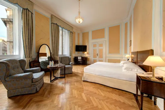A bedroom with a large bed, two armchairs, a round side table, an oval dressing mirror, and a small desk and television in the background.