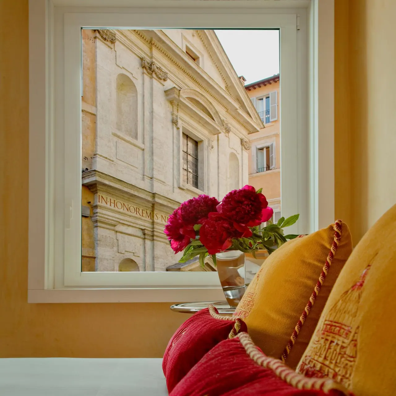 A window looks out onto a stone baroque church facade bearing an inscription, with red peonies in a glass vase and red and gold cushions in the foreground.